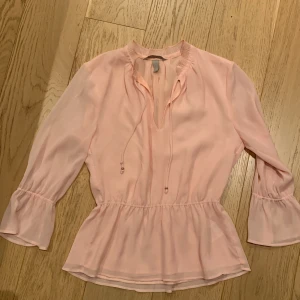 Ljusrosa blus från H&M - Säljer en superfin ljusrosa blus från H&M i storlek 34. Blusen har trekvartsärmar med en liten volangdetalj vid ärmsluten och en söt knytning vid halsen. Perfekt för vår och sommar! 💖