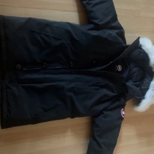 Svart vinterjacka från Canada Goose - Säljer en svart äkta vinterjacka från Canada Goose i storlek L. Jackan har en mysig pälskant på huvan och är perfekt för kalla vinterdagar. Den är i nyskick och har både dragkedja och knappar för extra skydd mot kylan. Supervarm Skick 10/10 