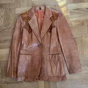 Läderjacka - Brun äderjacka i 100% genuine lether och foder i siden. Den har en slit baktill och en innerficka utöver de vanliga fickorna. Supersnygga detaljer, perfekt till hösten! Hittar ej storlek men passar mig som har S