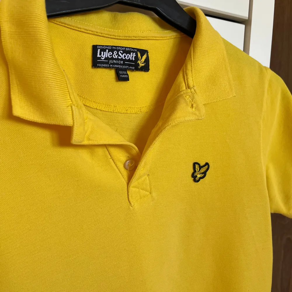 Säljer en snygg gul pikétröja från Lyle & Scott i storlek 12/13 år. Tröjan har en klassisk krage med knappar och Lyle & Scott-logotypen broderad på bröstet. Perfekt för sommardagar eller en avslappnad stil. Skicket är toppen, så passa på!. T-shirts.