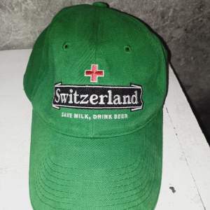 Säljer en grön keps med texten 'Switzerland' och 'Save Milk, Drink Beer' broderad på framsidan. Kepsen har en röd korsdetalj ovanför texten. Perfekt för sommarens äventyr eller som en rolig accessoar!