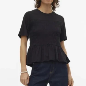 Svart topp med volangdetalj - Snygg svart topp med korta ärmar och volangdetalj i nederkant. Toppen har en smockad design som ger en fin struktur och passform. Perfekt för en stilren look.