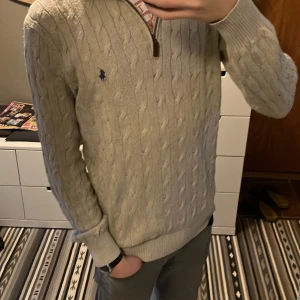  kabelstickad tröja från Ralph Lauren - Säljer en stilren beige kabelstickad tröja från Ralph Lauren. Tröjan har en halv dragkedja och det ikoniska blå logotypbroderiet på bröstet. Perfekt för kyligare dagar med sin mjuka och bekväma passform.