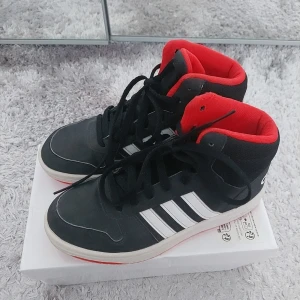 Svarta Adidas sneakers med röd insida - Snygga svarta Adidas sneakers med klassiska vita ränder och en cool röd insida. Skorna har snörning och en bekväm passform, perfekt för en sportig look. Adidas-loggan syns tydligt på baksidan.