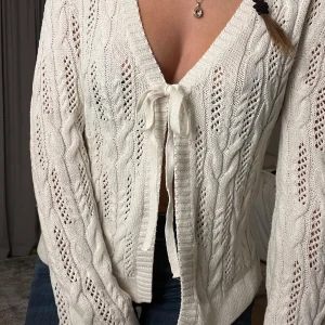 Vit stickad kofta - Säljer en vit stickad kofta med kabelstickning och knyt framtill. Koftan har långa ärmar och en djup v-ringning, passar fint med ett linne under också