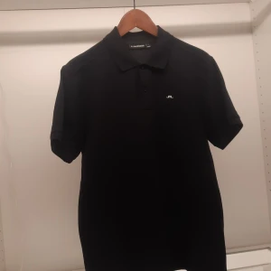 Svart pikétröja från J.Lindeberg - Snygg svart pikétröja från J.Lindeberg med klassisk krage och knappar framtill. Tröjan har en liten vit logotyp på bröstet och är perfekt för en stilren look.