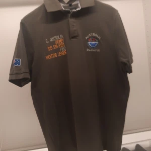 Olivgrön pikétröja från Est. 1982 Sweden - Säljer en olivgrön pikétröja från Est. 1982 Sweden med broderade detaljer och text på bröstet och ryggen. Tröjan har en klassisk krage med randigt mönster på insidan och korta ärmar. Perfekt för en avslappnad stil.