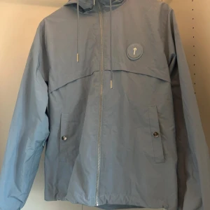 Ljusblå vindjacka från Trapstar - Säljer en ljusblå vindjacka från Trapstar med dragkedja och huva. Jackan har ett tryck med Trapstar-logga på ryggen och en rund logga på bröstet. Tillverkad i 100% polyester, perfekt för blåsiga dagar.