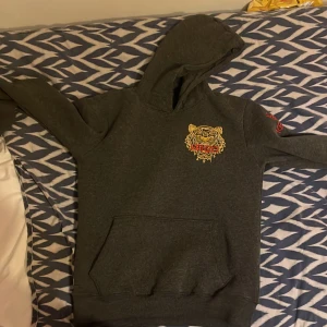 Grå hoodie från Kenzo - Säljer en snygg grå hoodie från Kenzo med ett coolt tigertryck på bröstet och broderad logga på ärmen. Tillverkad i 100% bomull, perfekt för en casual look. Den har en klassisk känguruficka och huva.