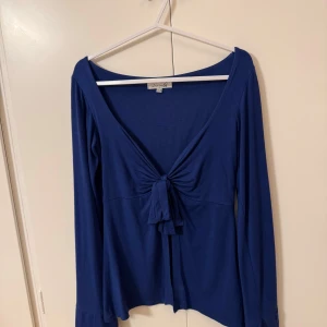 Blå långärmad topp från Design By Si - Snygg blå långärmad topp från Design By Si med knytning framtill. Toppen har en djup v-ringning och är tillverkad i ett mjukt material som ger en skön passform.
