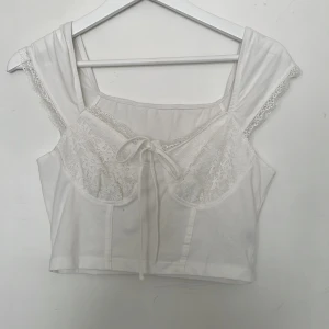 Vit spetsprydd topp - Säljer en söt vit topp med spetsdetaljer och knytning framtill. Toppen har breda axelband och en vacker spetskant som ger en romantisk känsla. Perfekt för sommarens varma dagar.