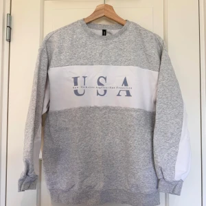 Grå och vit sweatshirt med USA-tryck - Säljer en grå och vit sweatshirt med stort USA-tryck på framsidan. Tröjan har långa ärmar och en rund halsringning. Perfekt för en avslappnad stil. Hör av dig vid intresse!