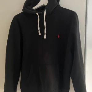 Svart hoodie från Polo Ralph Lauren - Säljer en klassisk svart hoodie från Polo Ralph Lauren med det ikoniska röda logotypen på bröstet. Tröjan har en justerbar huva med vita snören och en praktisk magficka. Perfekt för en stilren och bekväm look. ‼️PRIS GÅR ATT DISKUTERAS ‼️