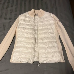 Vit dunjacka från Moncler - Säljer en stilren vit dunjacka från Moncler med quiltad design och dragkedja framtill. Jackan har långa ärmar och en ribbad krage för extra komfort. Perfekt för kyligare dagar och ger en lyxig känsla. Moncler-logga på ärmen ger en exklusiv touch.