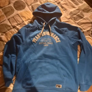 Blå hoodie från Peak Performance - Säljer en blå hoodie från Peak Performance med vit text på bröstet. Den har en klassisk känguruficka och justerbar huva med vita snören. Perfekt för en avslappnad stil.