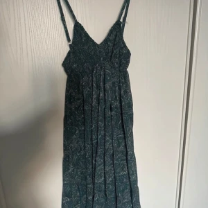 Grön mönstrad klänning  - Snygg grön klänning med paisleymönster från Meet Me There. Klänningen har smala axelband och en v-ringad design som ger en elegant look. Perfekt för varma dagar med sitt lätta och luftiga material.