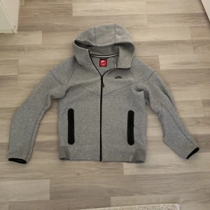 Grå Nike tech Fleece - Säljer en grå hoodie från Nike med dragkedja och huva. Jackan har svarta detaljer vid fickorna och ärmsluten. Perfekt för en sportig look eller avslappnade dagar.