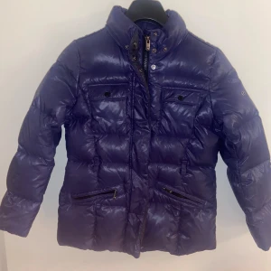 dunjacka från Eddie Pen - Snygg lila dunjacka från Eddie Pen med dragkedja och knappar. Jackan har långa ärmar och praktiska fickor med dragkedja. Perfekt för kyliga dagar med sin quiltade design. Stl 10 ca140 blålila