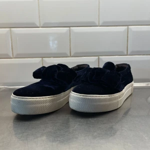 Mörkblå sammetssneakers med rosett från billi bi - Snygga mörkblå slip in sneakers i sammet med en stor dekorativ rosett på ovansidan. Skorna har en vit, tjock sula i storlek 40-41.  Skriv privat ifall ni vill ha fler bilder på skorna. Kan mötas upp i Varberg💕 pris går att diskutera