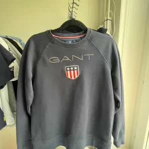 Mörkblå sweatshirt från GANT - Säljer en stilren mörkblå sweatshirt från GANT med broderad logga och emblem på bröstet. Tröjan har långa ärmar och rund halsringning. Perfekt för en avslappnad och snygg look. Pris kan diskuteras!