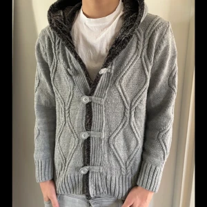 Fodrad kofta - Mysig grå stickad kofta med knappar och en huva fodrad med mjuk päls. Perfekt för kyliga dagar. Den har ett snyggt kabelstickat mönster och ribbade muddar.