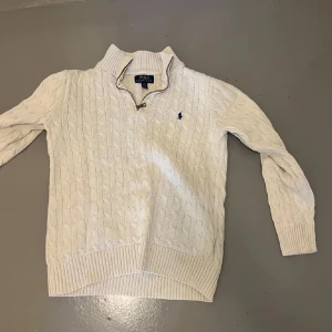 Vit kabelstickad tröja från Ralph Lauren - Säljer en stilren vit kabelstickad tröja från Ralph Lauren med en liten broderad logga på bröstet. Tröjan har en halv dragkedja vid halsen och ribbstickade muddar vid ärmslut och nederkant. Perfekt för kyligare dagar.