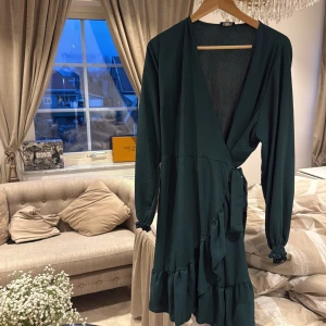 Mörkgrön omlottklänning  - Snygg mörkgrön omlottklänning från Zack London med långa ärmar och volangdetaljer. Klänningen har en knytning i midjan och en v-ringad halslinje, perfekt för en elegant look. Motsvarar XL