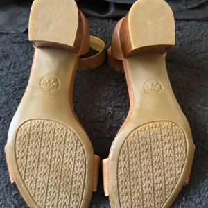 Lena sandaler från Michael Kors - Snygga bruna sandaler från Michael Kors i skinn med en bekväm klack. De har en stilren design med öppen tå och justerbart spänne runt ankeln. Perfekta för sommarens alla tillfällen.