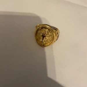 Guldfärgad ring med emblem - Elegant guldfärgad ring med ett detaljerat emblem på framsidan. Ringen har en rund form och är dekorerad med intrikata mönster runt emblemet. Perfekt för att ge en klassisk touch till din stil.