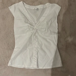 Vit topp från H&M - Säljer en stilren vit topp från H&M i storlek XS. Toppen har en v-ringad design med en knutdetalj framtill och korta ärmar. Perfekt för en enkel och fräsch look. säljes då den är lite liten för mig 
