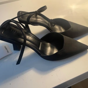 Svarta skor med klack och knytband - Svarta pumps från Pull&Bear med spetsig tå, klack och ett knytband runt ankeln. Perfekta för en kväll ute eller liknande! Supersnygga men tyvärr aldrig använda då jag  insett att jag inte kan gå i klackar!