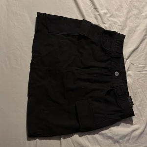Svart kjol med fickor - Snygg svart kjol med praktiska fickor på sidorna i jeans material och knappstängning fram. Perfekt för en casual look. Kjolen har en enkel och stilren design som gör den lätt att matcha med olika toppar. Säljer då den inte kommit till användning särskilt ofta.