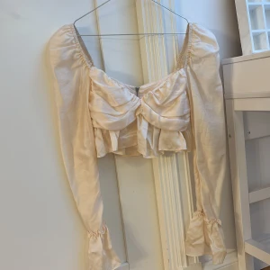 Vit blus med volangdetaljer - Säljer en cropped blus från H&M med volangdetaljer och långa ärmar. Blusen har en dragkedja i ryggen och en vacker rynkad design framtill. Är aldrig använd! Skriv om du har fler frågor!