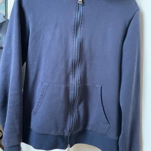 Mörkblå hoodie från Moncler - Säljer en snygg mörkblå hoodie från Moncler med dragkedja och fickor framtill. Hoodien har en röd och grå detalj i luvan och ribbade muddar vid ärmslut och nederkant. Perfekt för en avslappnad stil. Mindre hål på höger arm pris diskuteras i dm.