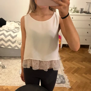 Vit ärmlös topp med spetsdetalj - Säljer en stilren vit ärmlös topp med vacker spetsdetalj längst ner. Perfekt för en somrig look eller att bära under en kavaj. Toppen har en lös passform och är gjord i ett lätt material.