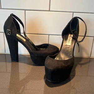 Svarta pumps från NLY Shoes - Snygga svarta pumps från NLY Shoes med hög klack och platåsula. Skorna har en justerbar rem runt ankeln och är tillverkade i mocka. Perfekta för att ge lite extra höjd och stil.