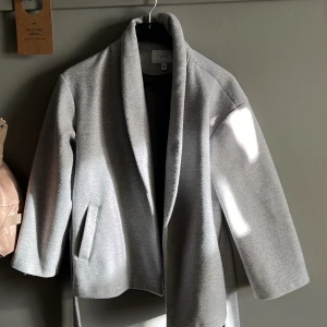 Grå kappa - Säljer en stilren grå kappa från vila. Den har en elegant design med bälte i midjan och långa ärmar. Perfekt för höst och vår. Materialet är mjukt och bekvämt. Den är två år gammal och är ganska nopprig. 