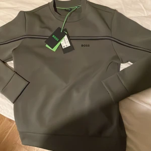 Grå sweatshirt från Boss - Snygg grå sweatshirt från Boss med diskret logga på bröstet. Tröjan har en stilren design med svarta linjer över bröstet och långa ärmar. Perfekt för en avslappnad och trendig look.