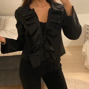 Svart kavaj med volangdetaljer - Snygg svart kavaj med volangdetaljer framtill. Den har långa ärmar och en elegant passform som passar perfekt för en stilren look. Perfekt för att ge en extra touch till din outfit. Storlek M men passar S