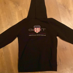 Svart hoodie från GANT - Säljer en stilren svart hoodie från GANT med deras klassiska logga och texten 'American Sportswear' på framsidan. Perfekt för en avslappnad look. Hoodien har långa ärmar och en bekväm huva. Ord pris 699kr