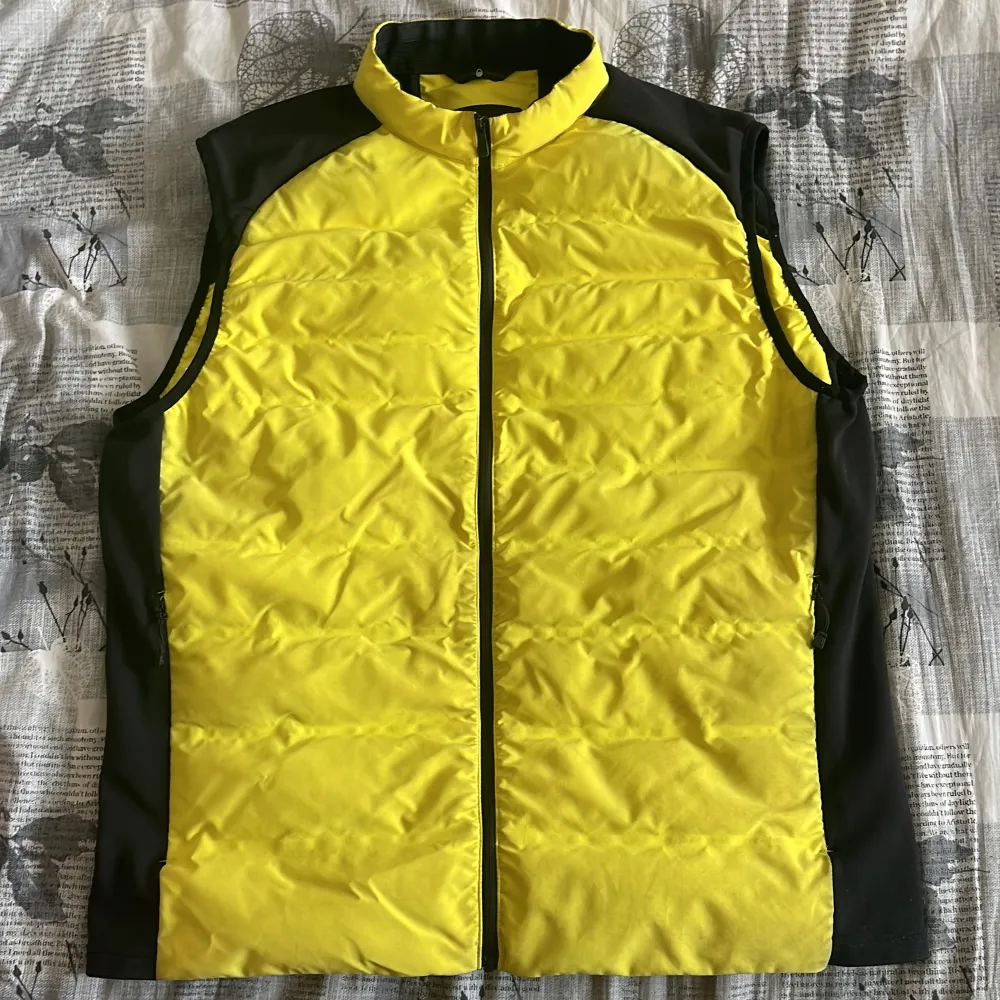Snygg gul och svart väst med dragkedja framtill. Perfekt för en sportig look eller som ett extra lager under kyliga dagar. Västen har en modern design med kontrasterande svarta paneler på sidorna.. Takit.