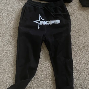 Svart tracksuit - Säljer en svart hoodie och matchande byxor med ett stort stjärntryck och texten 'NOFS' i vitt. Hoodien har en klassisk design med känguruficka och långa ärmar. Byxorna har en bekväm passform med resår i midjan och dragsko. Storlek L men skulle säga att den är liten i storleken, i alla fall byxorna. Oanvända!