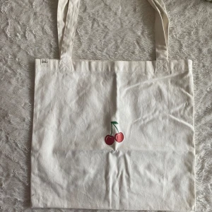 Vit shoppingväska med körsbärsmotiv - Säljer en söt vit totebag med ett körsbärsmotiv på framsidan. Väskan har långa handtag och är perfekt för att bära med sig det nödvändigaste. Enkel och stilren design som passar till många tillfällen. Har 2 små fläckar på baksidan som säkert går att få bort med tvätt. 