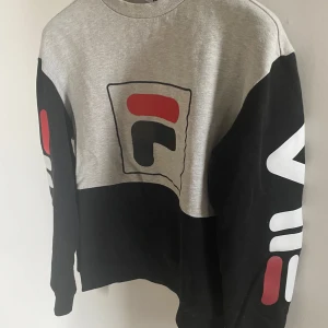Grå och svart tröja från FILA - Grå och svart tröja från FILA med deras ikoniska logga i rött och vitt på bröstet och ärmarna. Tröjan har långa ärmar och en bekväm passform, 
