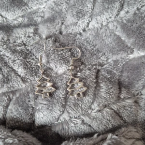 Silverfärgade örhängen med julgransmotiv - Snygga och festliga silverfärgade örhängen i form av julgranar. Perfekta för att ge en glittrig touch till din juloutfit. Lätta att bära med krokfäste.