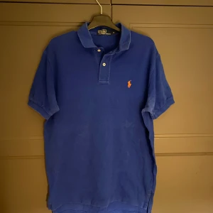 Blå pikétröja från Ralph Lauren - Säljer en klassisk blå pikétröja från Ralph Lauren med den ikoniska loggan broderad i rött på bröstet. Tröjan har korta ärmar och en stilren krage med knappar. Perfekt för en avslappnad men stilfull look.