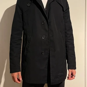 Svart kappa med knappar - Snygg svart kappa med knappar framtill. Den har en klassisk design med långa ärmar och en stilren look. Perfekt för höst och vår.