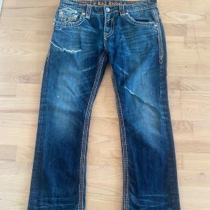 Blå jeans från Rock Revival - Snygga blå jeans från Rock Revival med slitna detaljer och broderade bakfickor. Jeansen har en unik design, priset går att diskuteras och skruv för fler bilder!🚁💓🤞