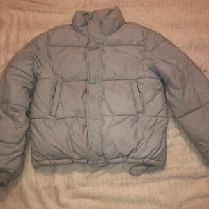 Baby blå pufferjacka - Säljer en snygg baby blå pufferjacka i bra skick. Jackan är perfekt för kalla dagar med sin vadderade design och höga krage. Den har långa ärmar och praktiska fickor framtill. Passar perfekt för både höst och vinter. Perfekt för dig som vill hålla stilen i kylan!