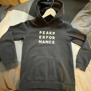 Svart hoodie från Peak Performance - Säljer en svart hoodie från Peak Performance i mycket bra skick. Den har en klassisk design med vit text på bröstet och en stor ficka framtill. Perfekt för både vardag och träning. Superbekväm och stilren!